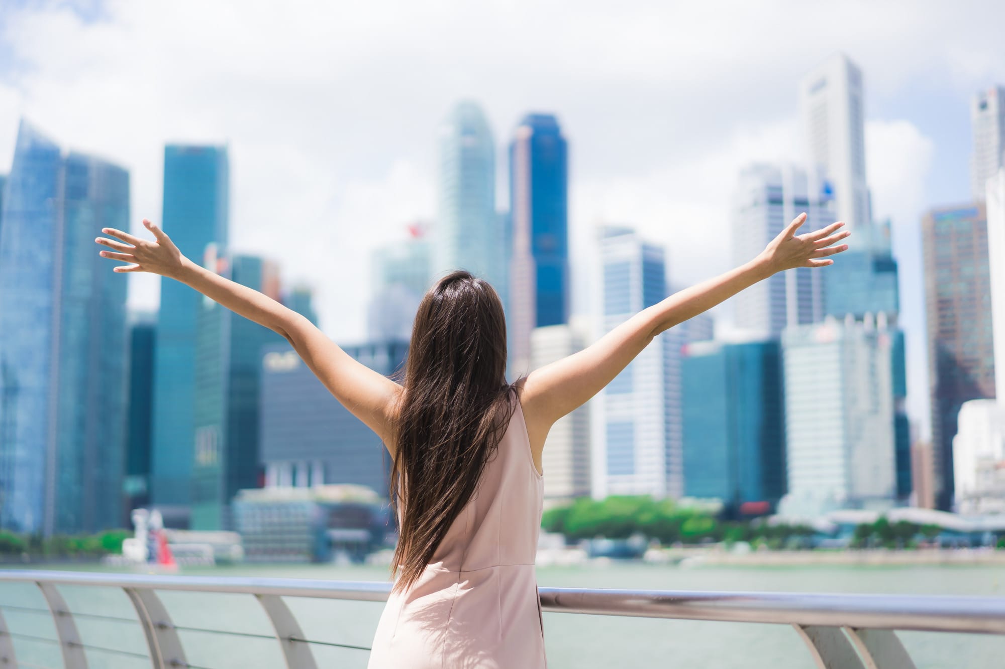 Woman with long hair raises both arms wide facing a blurred city skyline of tall glass skyscrapers.